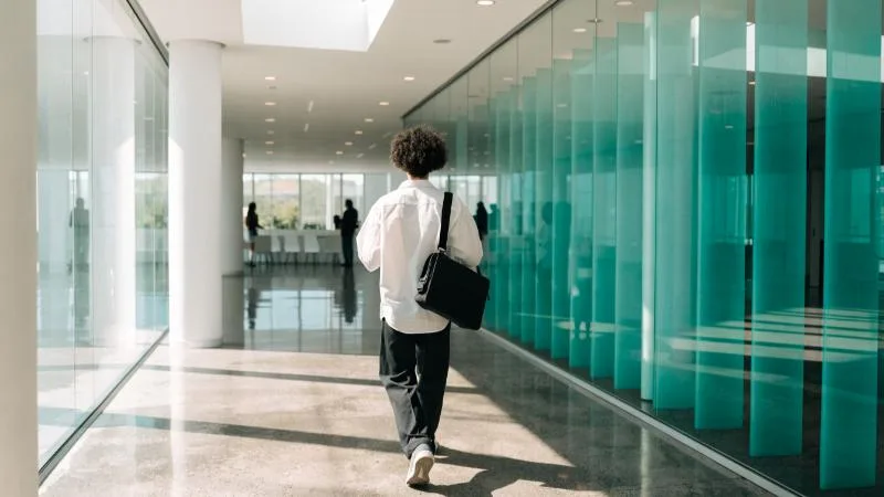 日差しの入るオフィスの廊下を、かばんを持って歩く若い社会人の後ろ姿